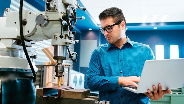 A young Latin engineer concentrated on calibrating a large drill with his laptop in his shop.