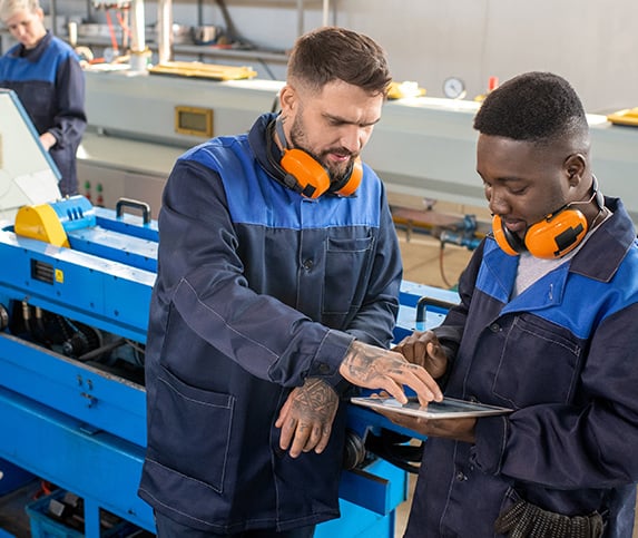 A black and white male workforce professional work through a manufacturing solution on the shop floor with a blue machine in the background.